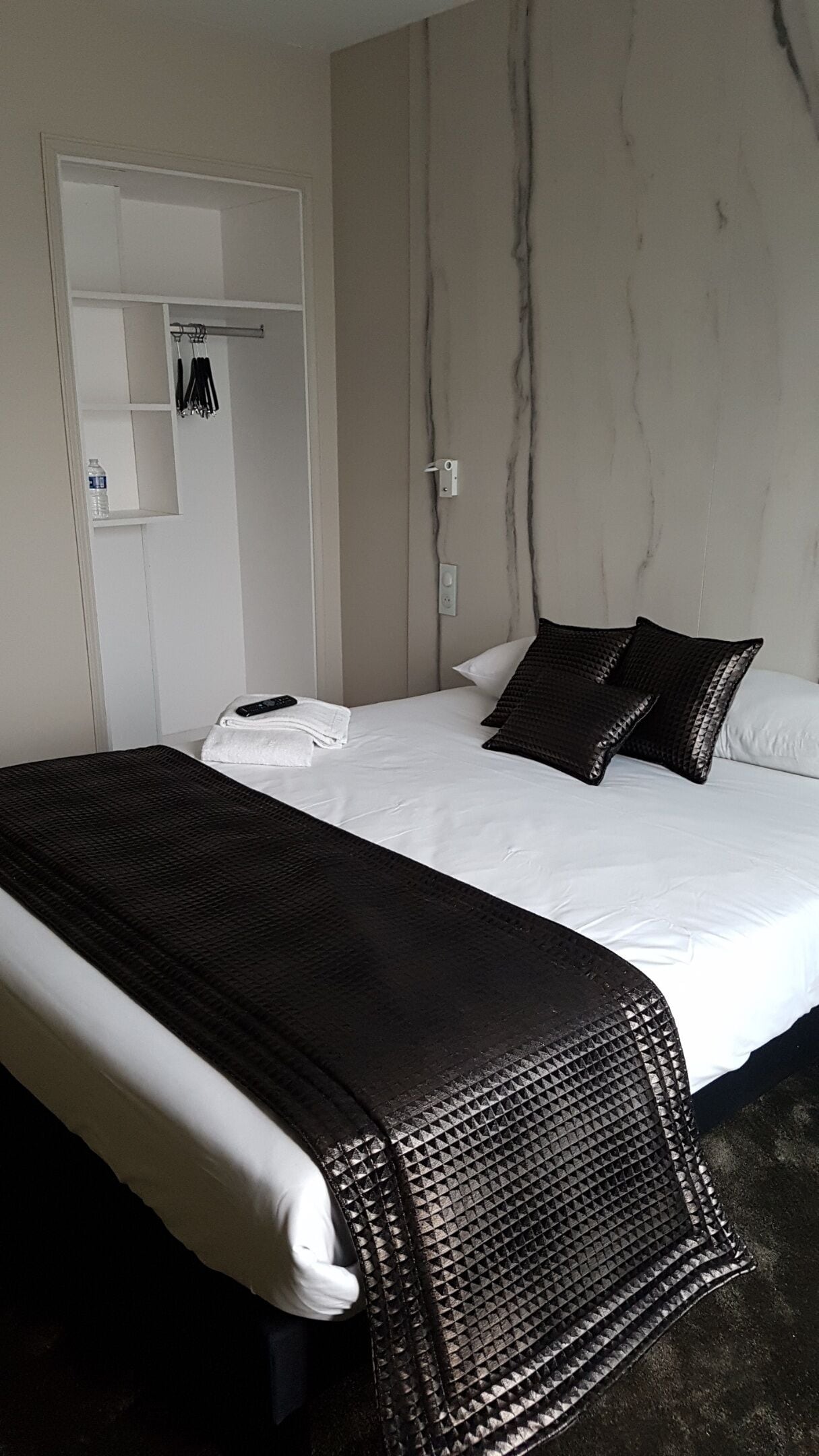 A hotel room with a bed, a white wall with a marble-like texture, a white door with a towel rack, and a black textured throw blanket on the bed.