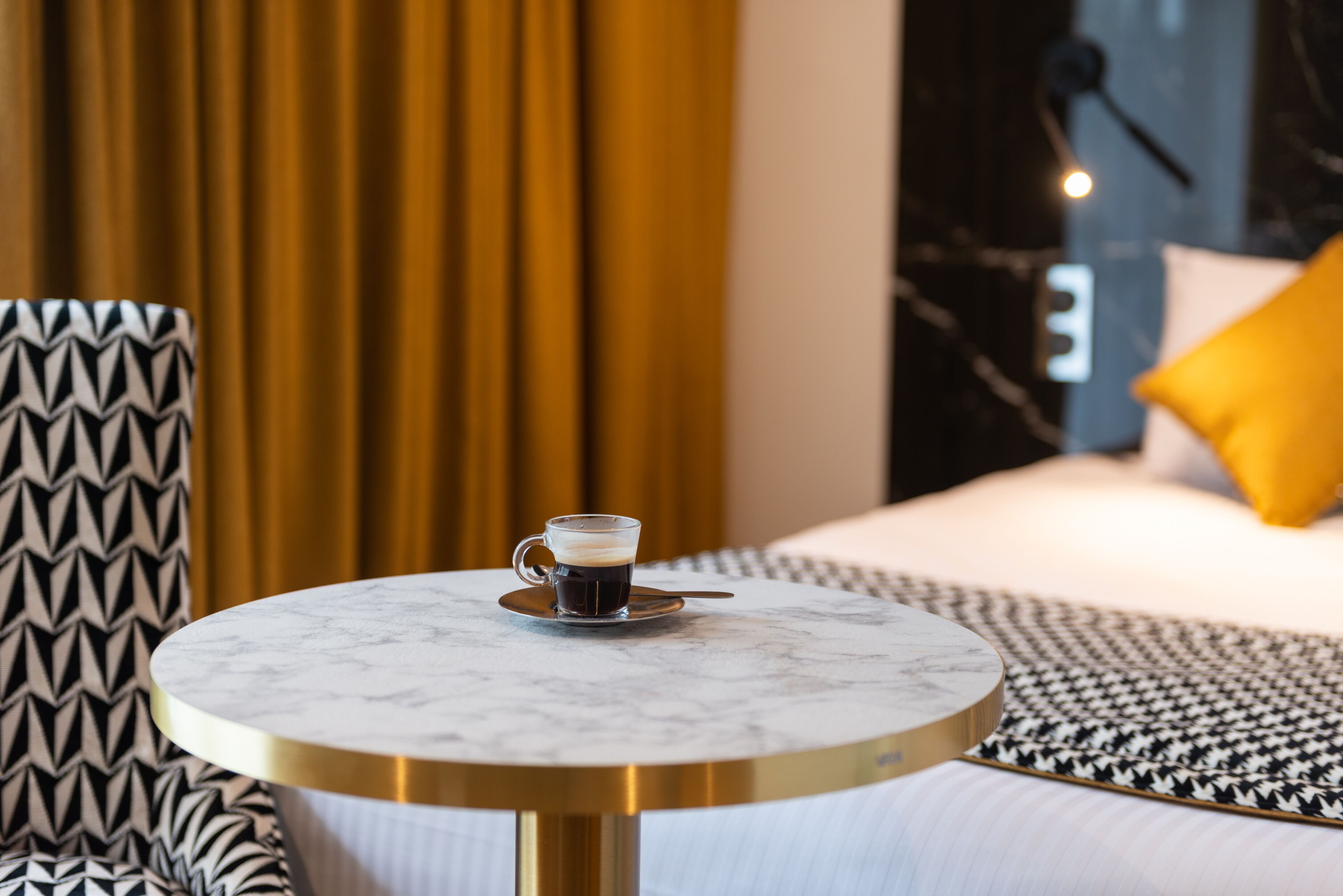 A round table with a coffee cup, a patterned chair, a bed with a yellow pillow, and a curtain in the background.