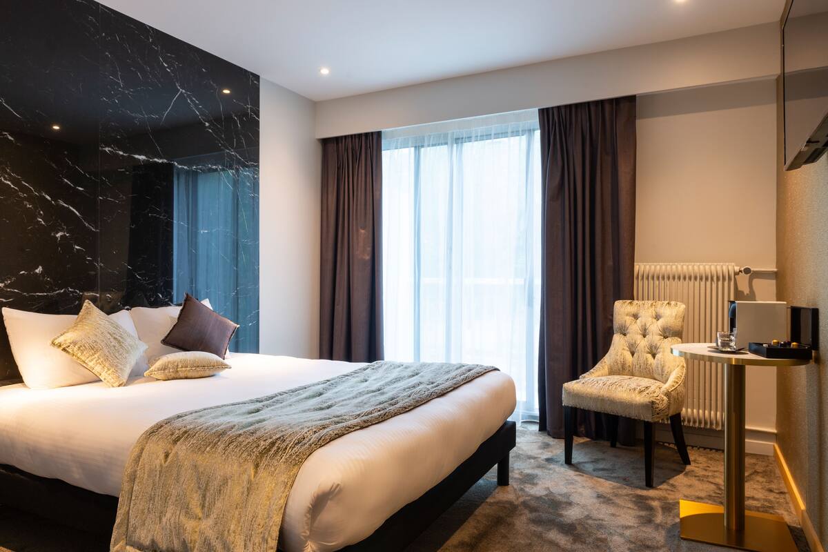 Habitación de hotel con una cama grande, una silla, una mesita y una ventana con cortinas.