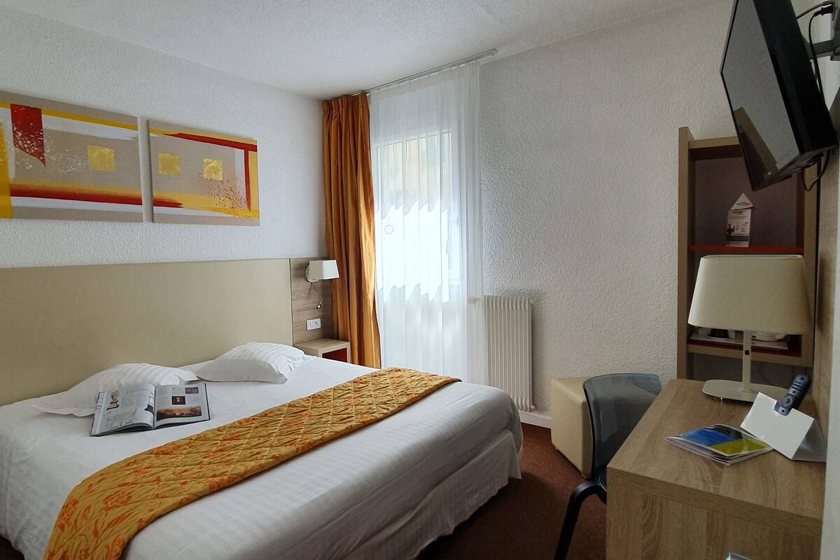 Habitación de hotel con cama, escritorio, silla, lámpara, televisor y ventana con cortinas.