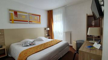 Habitación de hotel con cama, escritorio, silla, lámpara, televisor y ventana con cortinas.