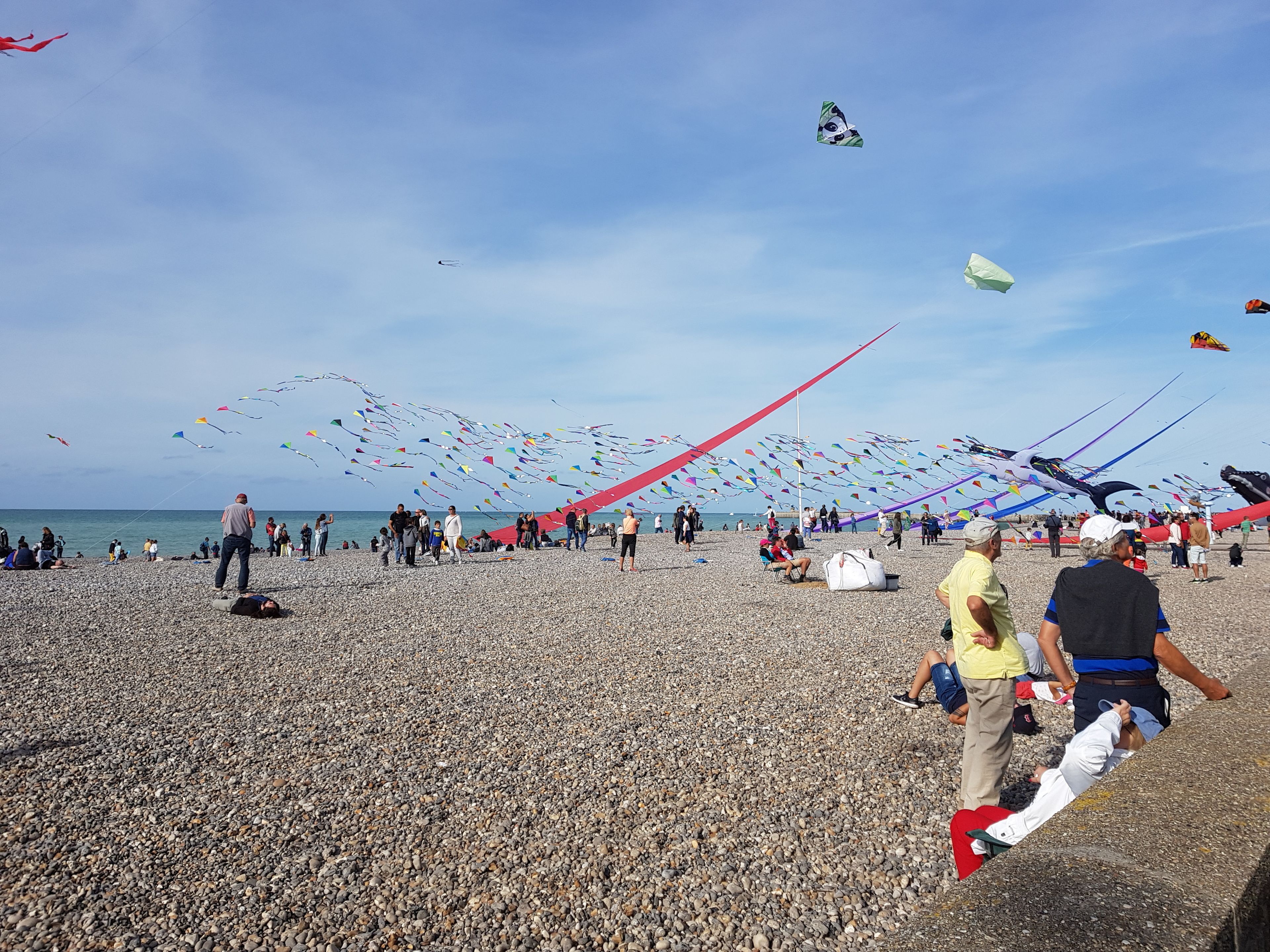 Une plage de galets où l’on voit des gens faire voler des cerfs-volants, sous un ciel dégagé.
