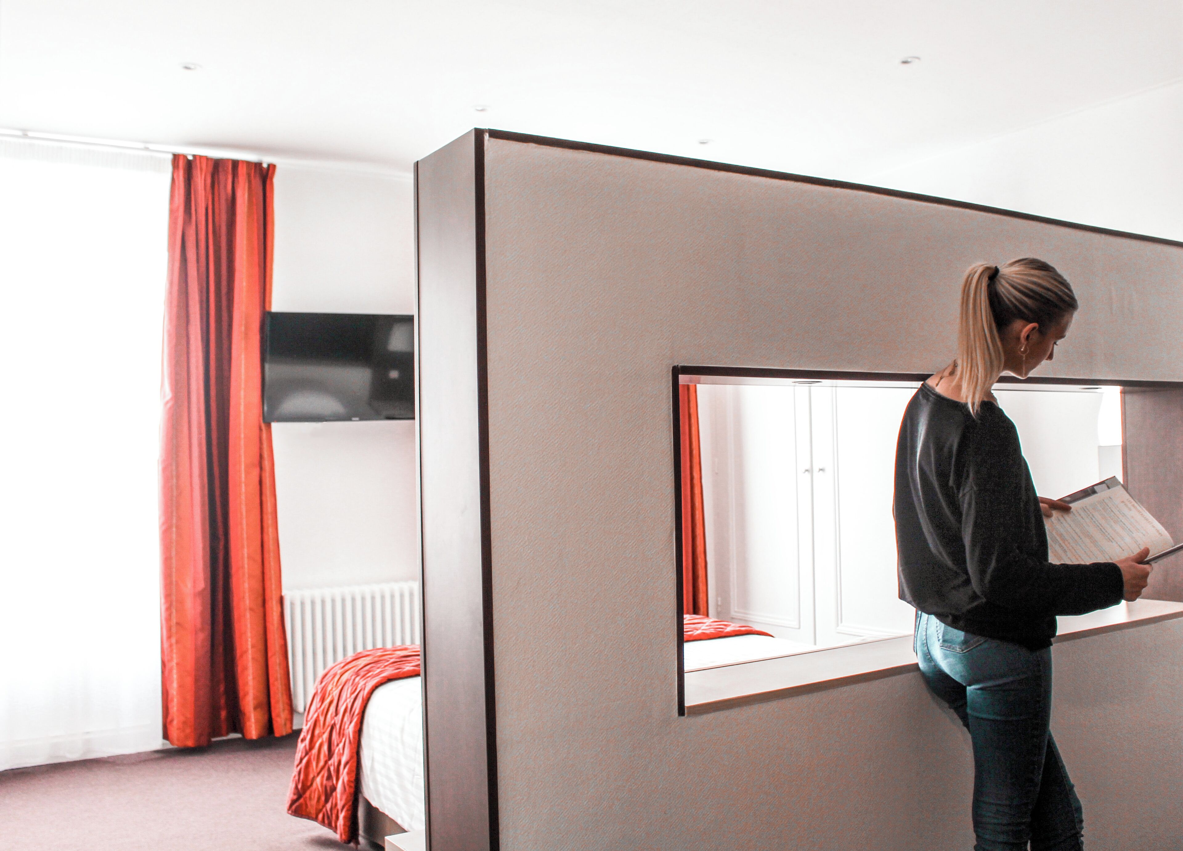 A modern hotel room with a large window, a bed with red curtains, and a person reading a book.
