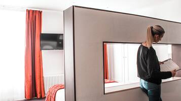 A modern hotel room with a large window, a bed with red curtains, and a person reading a book.