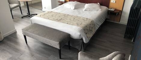 Habitación de hotel con cama, banca, silla, mesita y mesita de noche con lámpara.