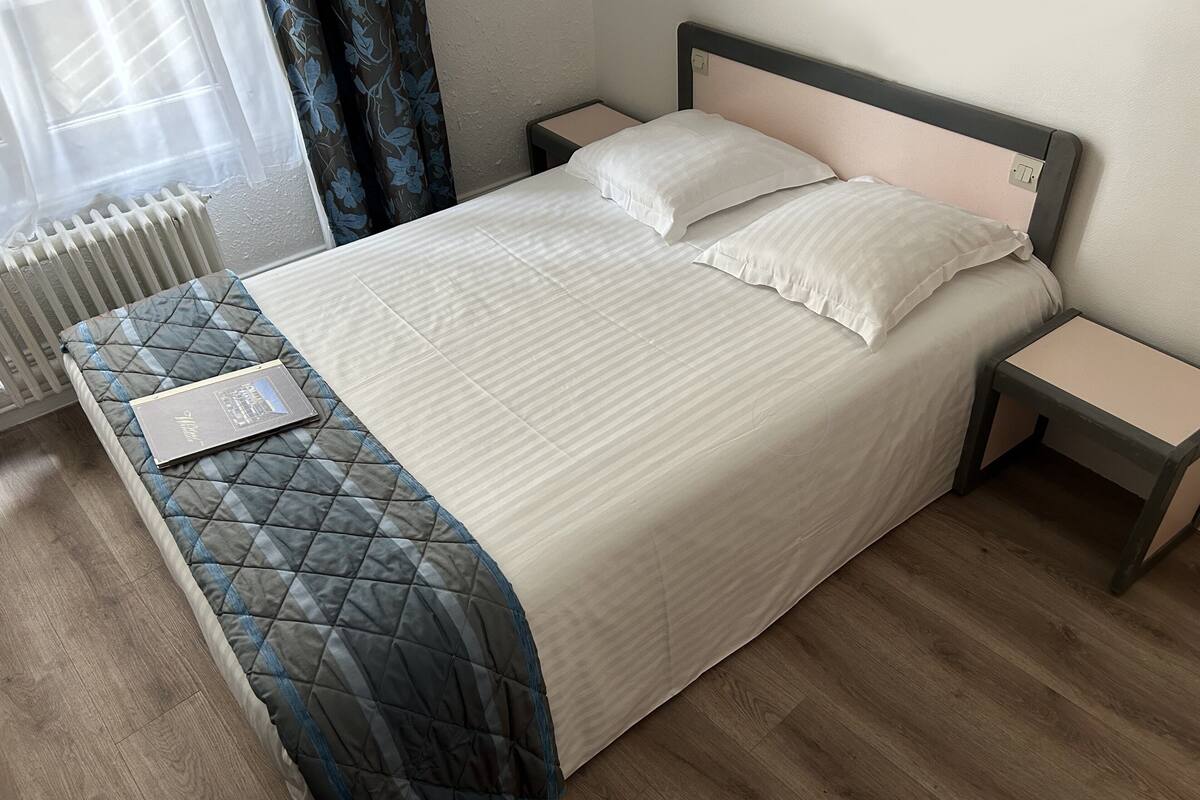 Una cama bien hecha con cubrecama acolchado, dos mesitas de noche, una lámpara de noche y una ventana con cortinas.