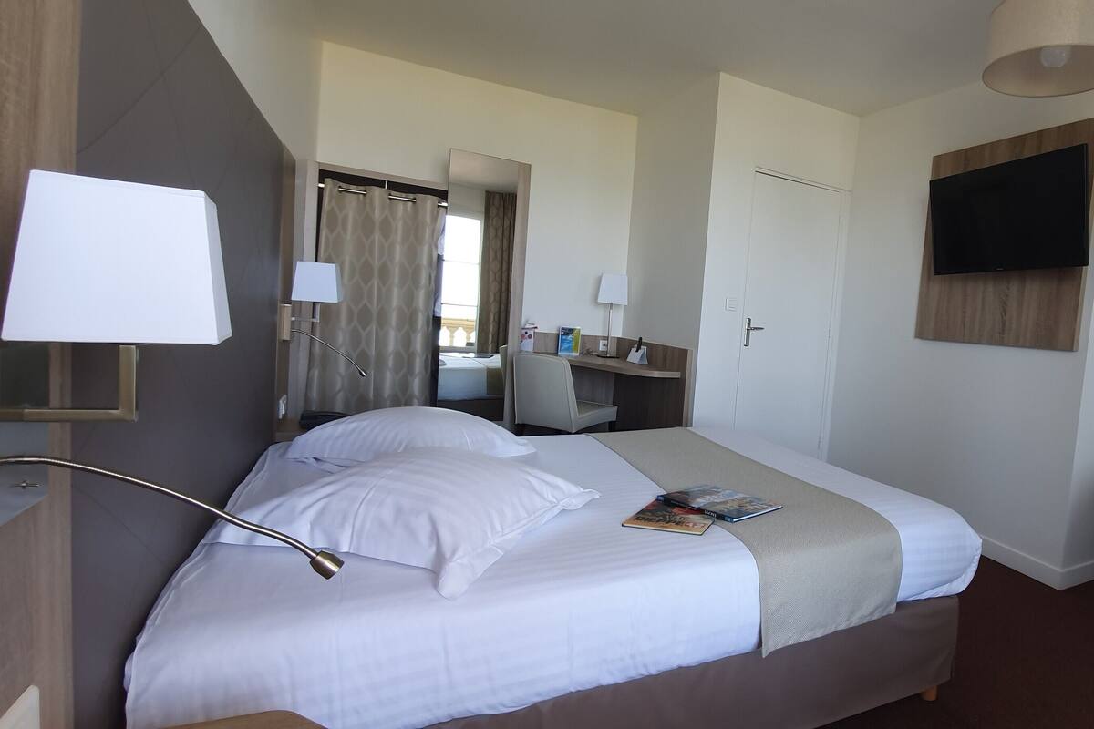 Habitación de hotel con cama, lámparas de noche, escritorio, silla, televisor y ventana con cortinas.