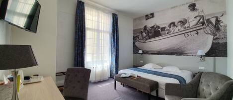 Habitación de hotel con cama, dos sillones, escritorio con lámpara, televisor y un mural grande con un barco.