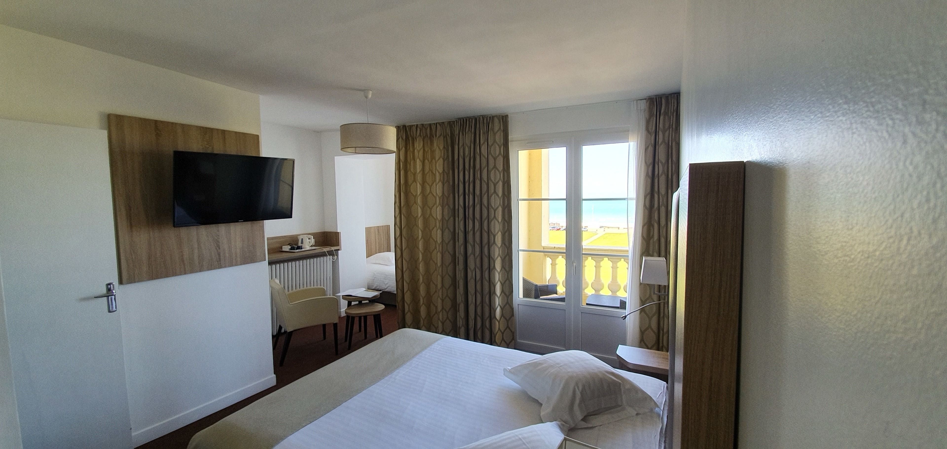 A hotel room with a bed, a TV mounted on the wall, a desk with a chair, a window with curtains, and a view of the outside.