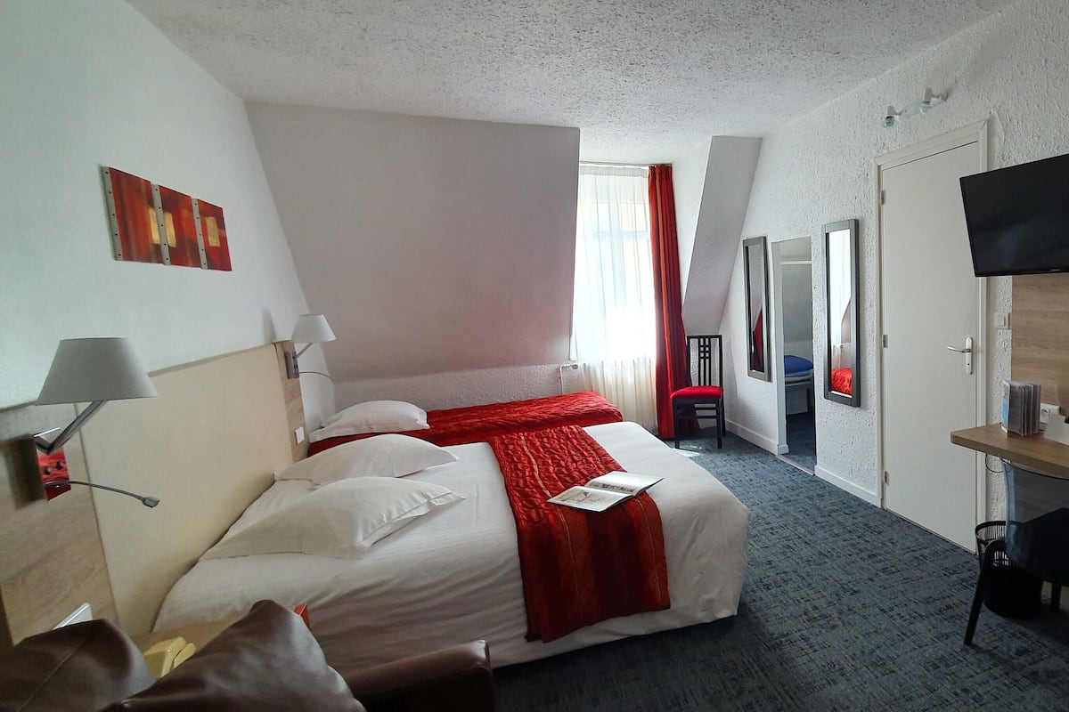 Una habitación de hotel con cama, escritorio, televisor, espejo y una silla roja.