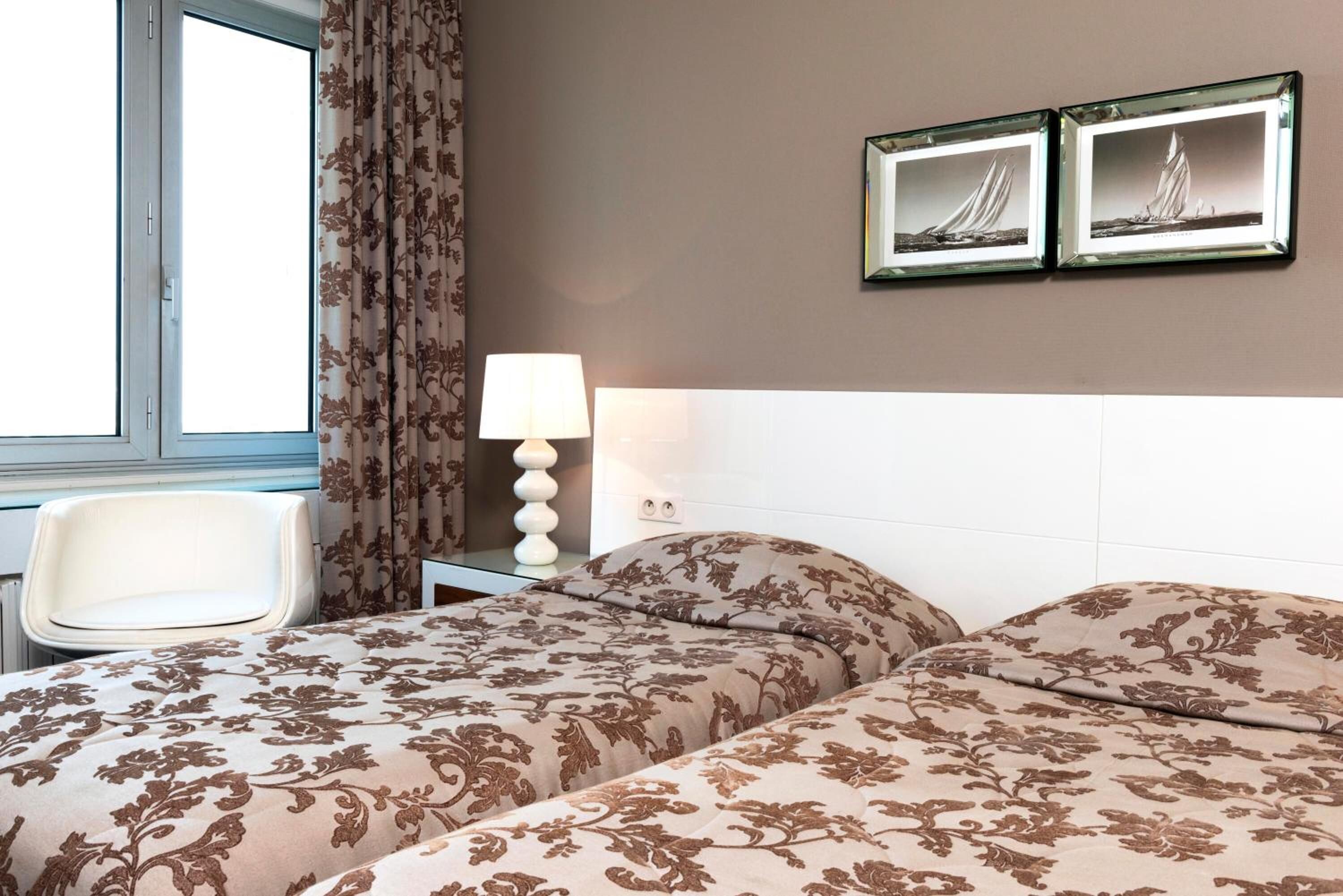 A hotel room with two beds, a chair, a nightstand with a lamp, and two framed pictures on the wall.