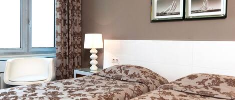 A hotel room with two beds, a chair, a nightstand with a lamp, and two framed pictures on the wall.