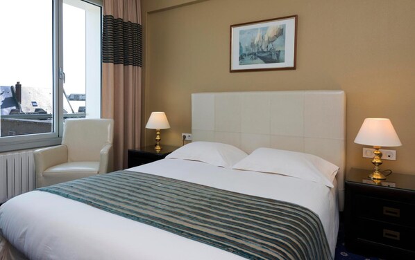 A hotel room with a bed, two bedside lamps, a chair, a window with curtains, and a painting on the wall.