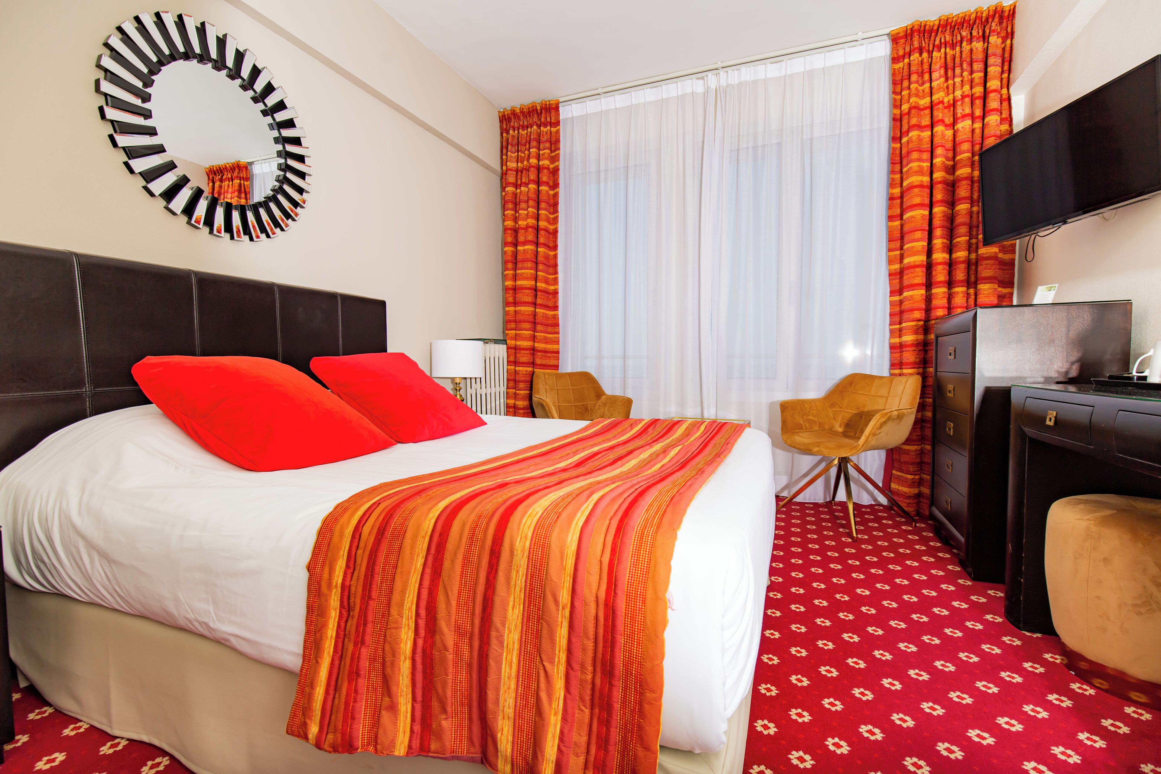 A hotel room with a bed, two chairs, a desk, a TV, and a large mirror on the wall.