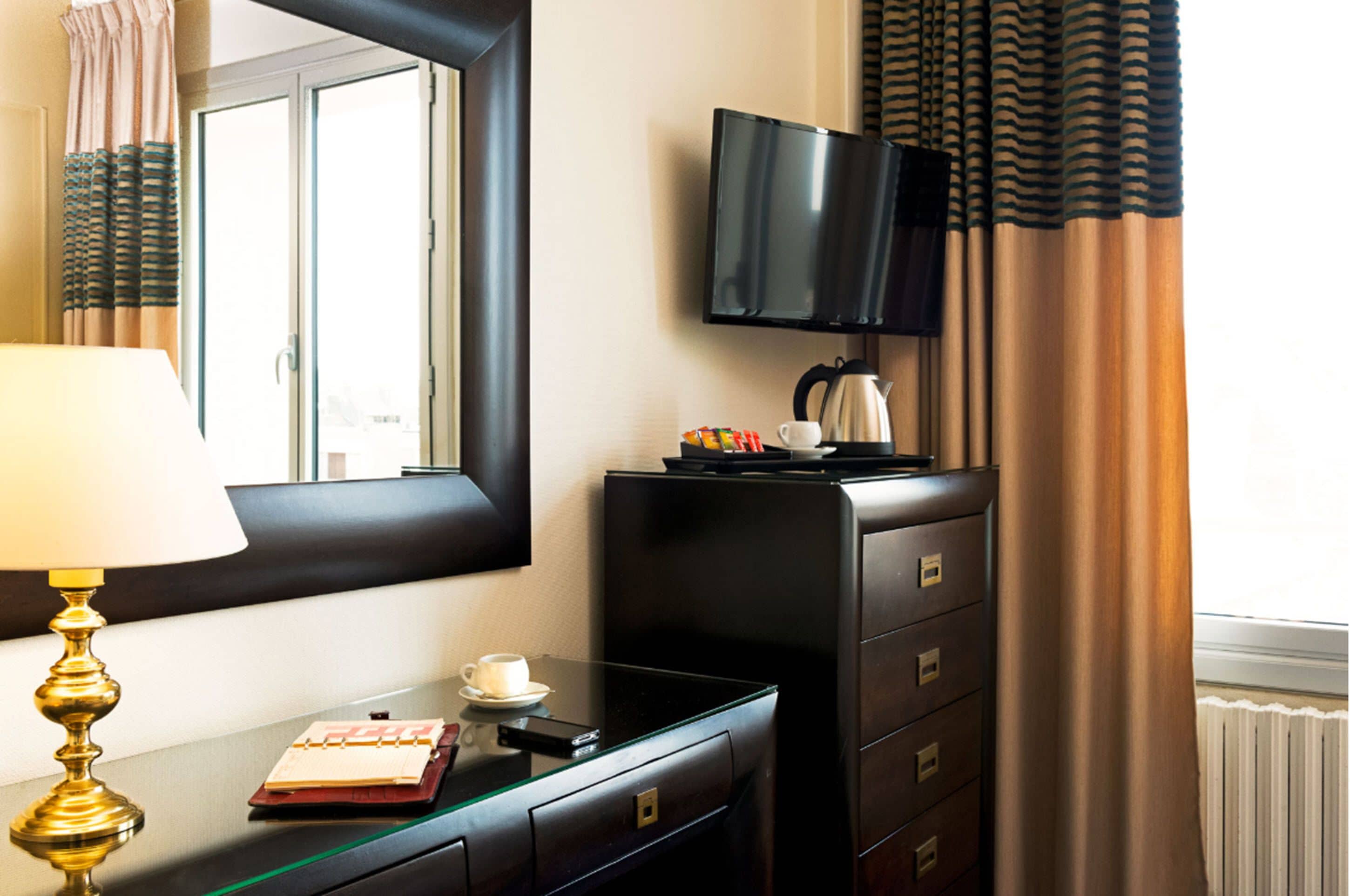 A hotel room with a large mirror, a flat-screen TV mounted on the wall, a coffee maker, a desk with a lamp, a cup, and a notepad.