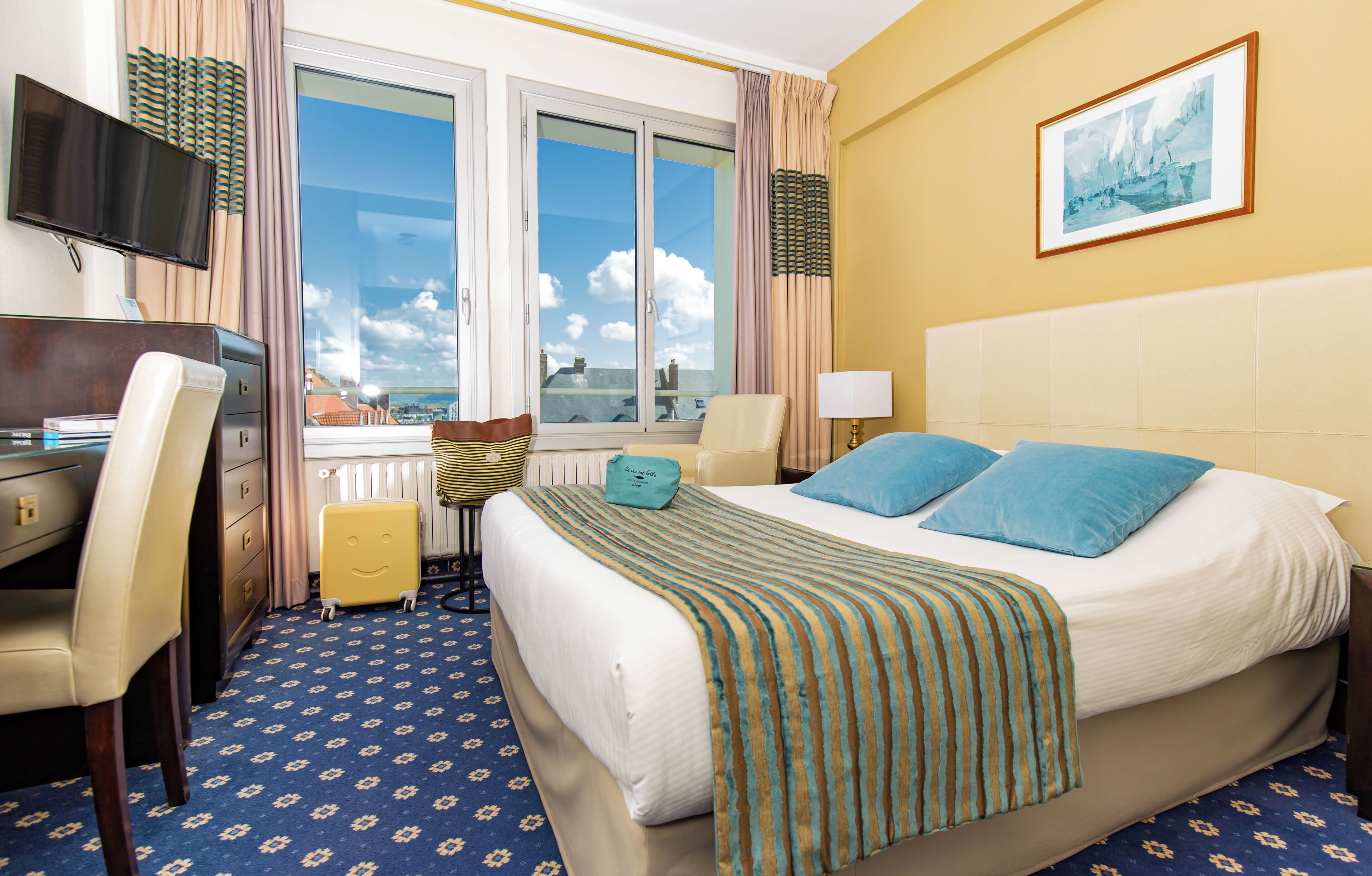 A hotel room with a large bed, a desk, a chair, a TV, and a window with a city view.