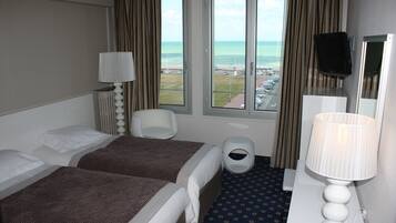 A hotel room with two beds, a desk with a lamp, a chair, a trash bin, and a large window offering a view of the beach and parking lot.