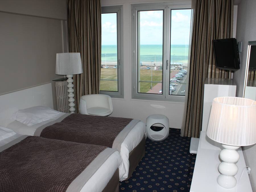 A hotel room with two beds, a desk with a lamp, a chair, a trash bin, and a large window offering a view of the beach and parking lot.