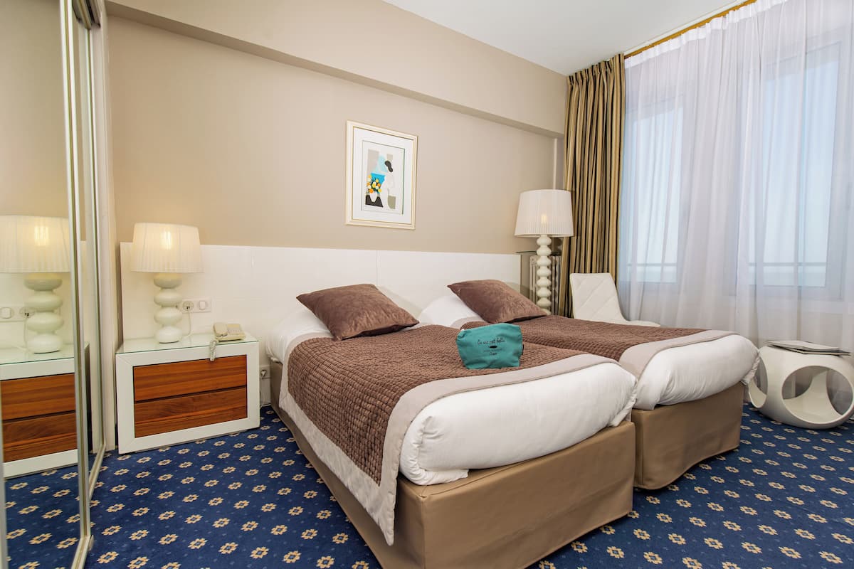 A hotel room with two beds, a bedside table with a lamp, a small bedside table with a phone, a wall-mounted picture, and a window with curtains.