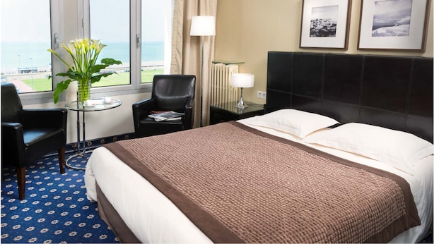 A hotel room with a bed, a chair, a small table with a vase of flowers, a lamp, and two framed pictures on the wall. - Hotel Aguado (Dieppe)