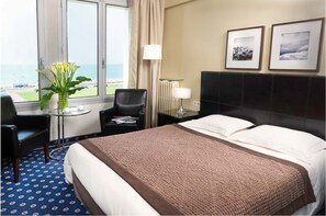 A hotel room with a bed, a chair, a small table with a vase of flowers, a lamp, and two framed pictures on the wall. - Hotel Aguado (Dieppe)