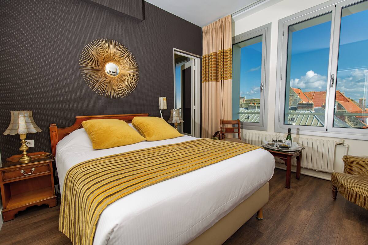 A hotel room with a large bed, a nightstand with a lamp, a chair, a table with a bottle, and a view of buildings through the window.