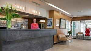 A reception area with a receptionist, a reception desk, a chair, and a large vase of flowers. - Hotel Aguado (Dieppe)