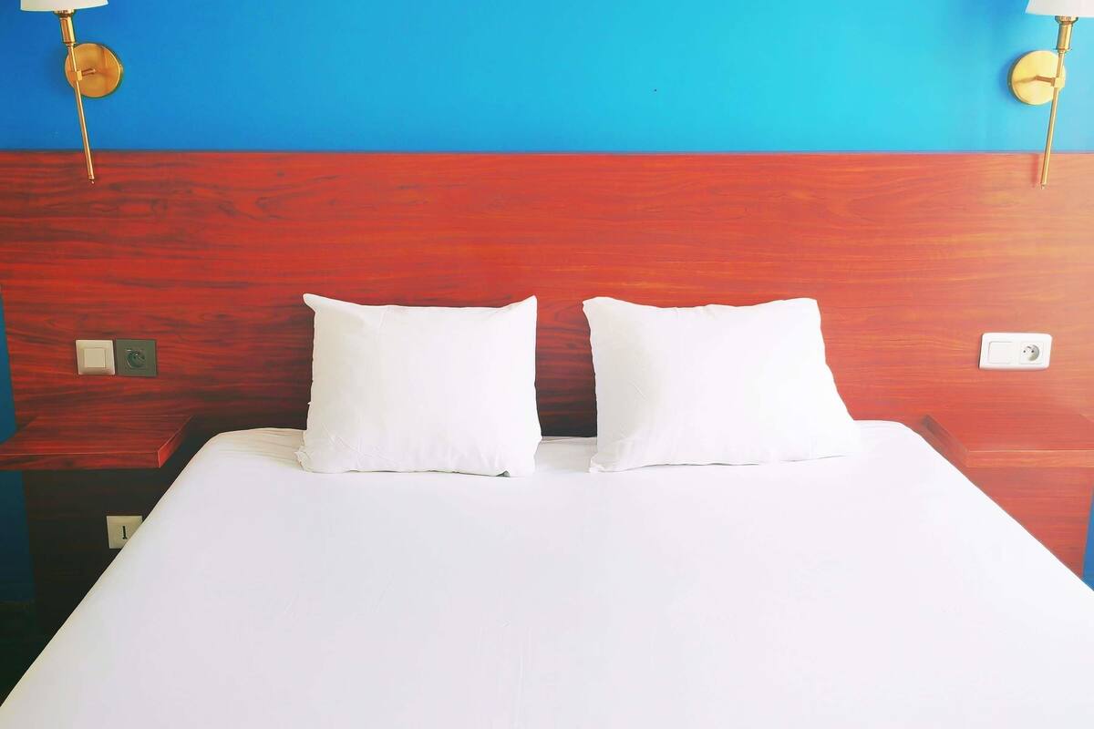 Habitación de hotel con cabecera de madera, dos almohadas blancas, una colcha blanca y dos lámparas fijadas a la pared.
