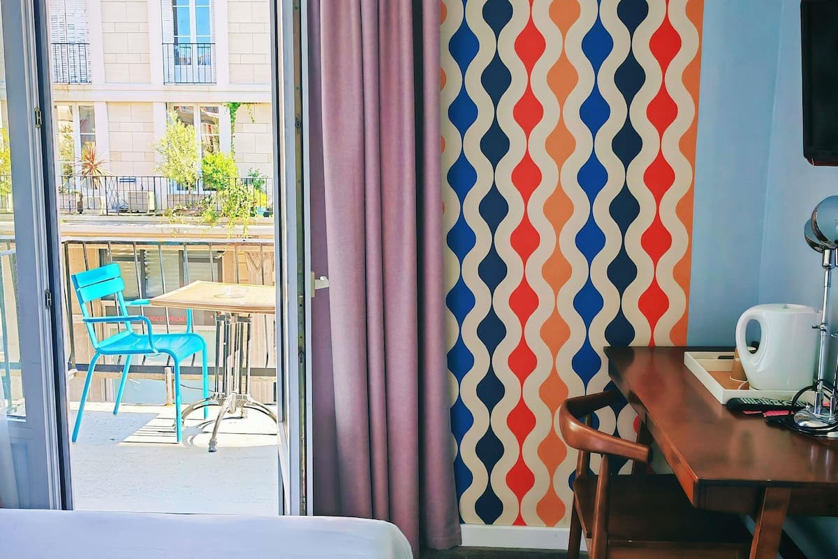 A hotel room with a bed, a desk, a chair, a table, a coffee maker, and a view of the outside.