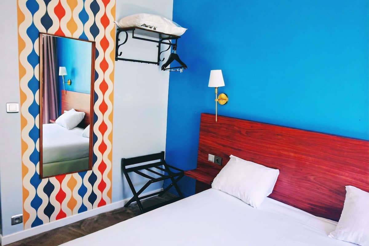 Habitación de hotel con cama, cabecera de madera, espejo, repisa en la pared y mesita de noche con lámpara.