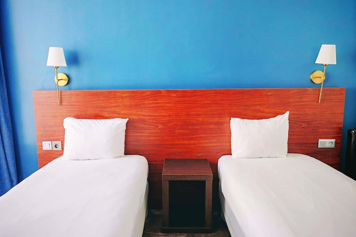 Habitación de hotel con dos camas individuales, cabecera de madera, lámparas de noche y mesitas auxiliares.