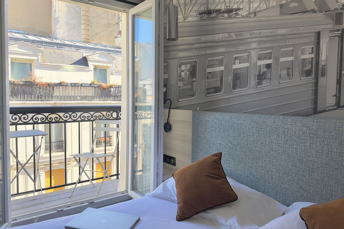 A hotel room with a bed, a laptop, a window with a view of a train, and a balcony with a railing.