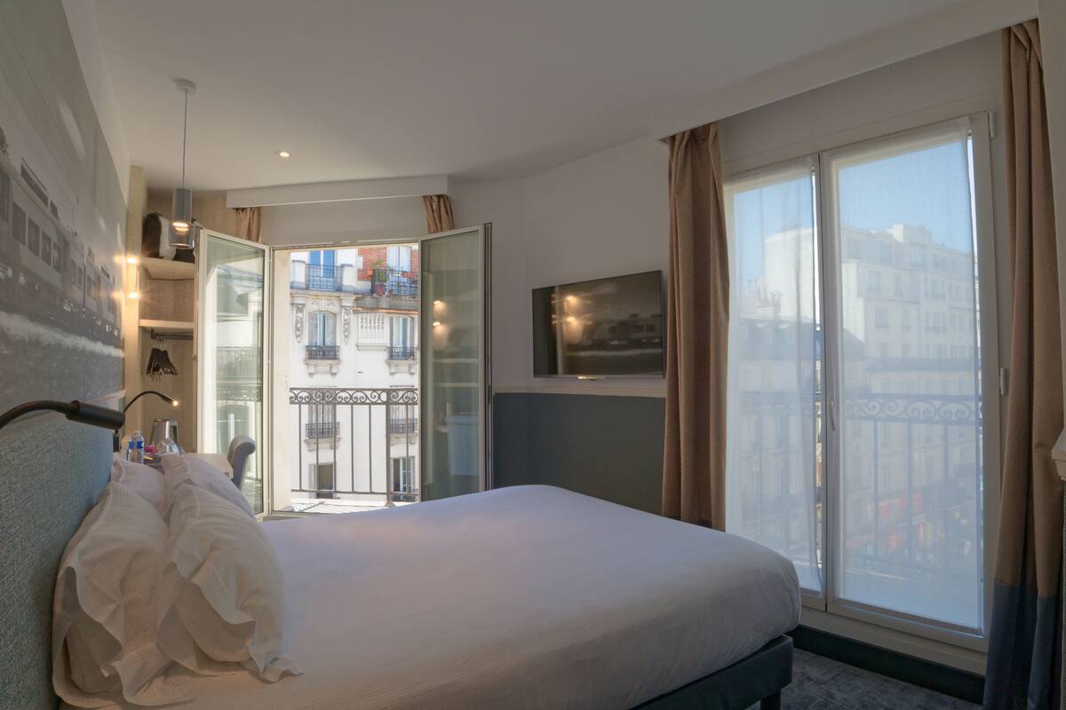 Ein Hotelzimmer mit einem großen Bett, einer Glastür, die auf einen Balkon führt, und Blick auf die Stadt.