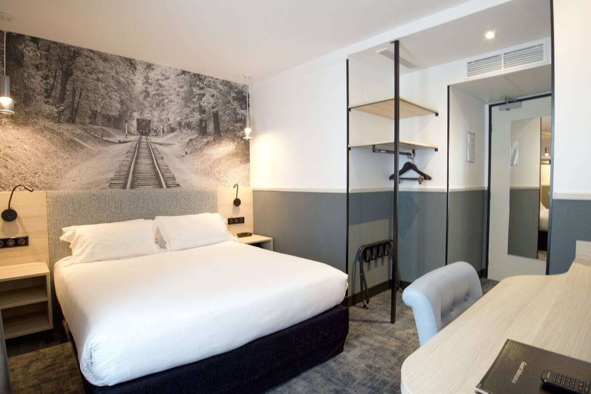 Ein Hotelzimmer mit einem großen Bett, einem Schreibtisch, einem Stuhl, einem Kleiderständer und einem Spiegel.