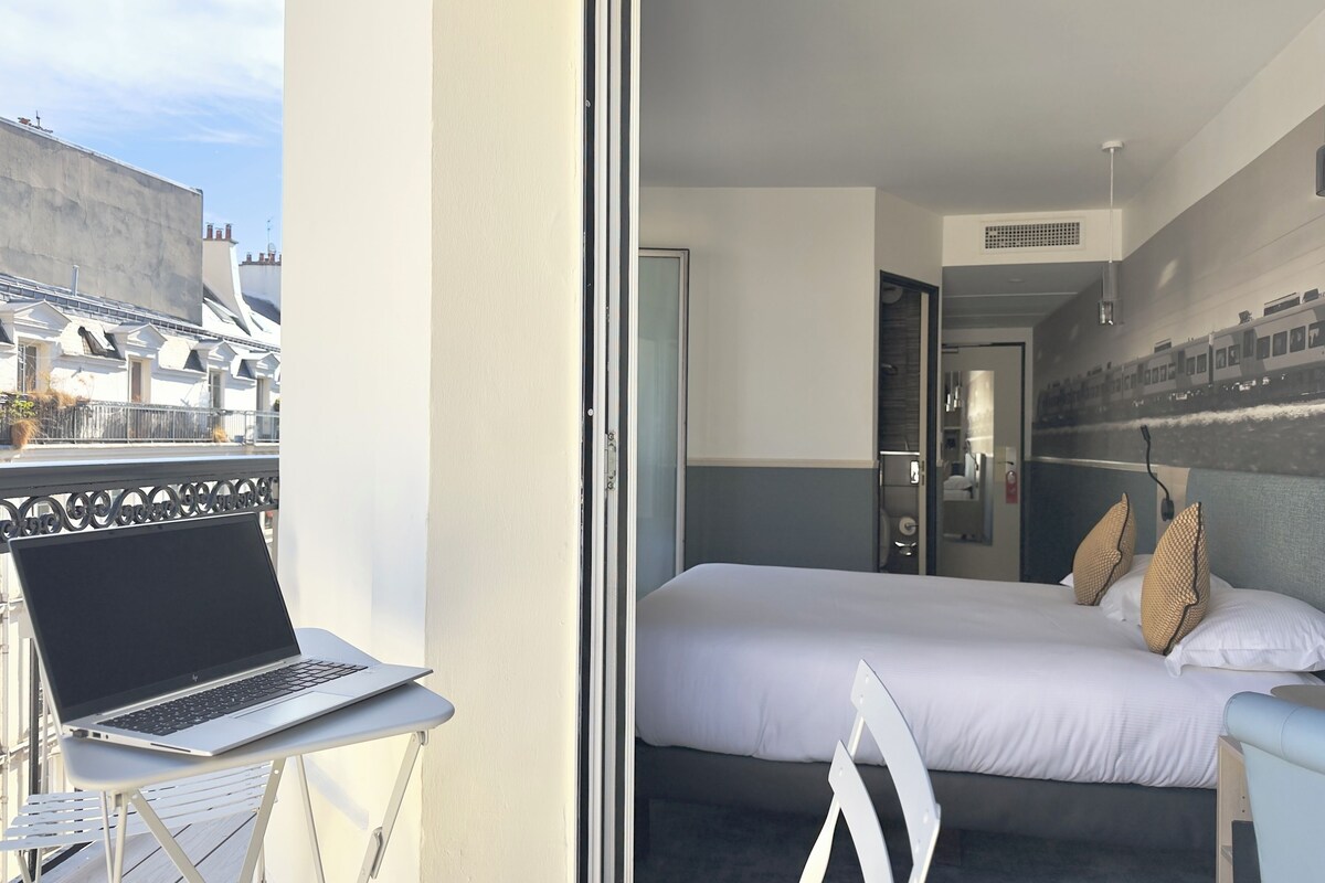 A hotel room with a bed, a laptop on a small table, and a view of the cityscape through the window.