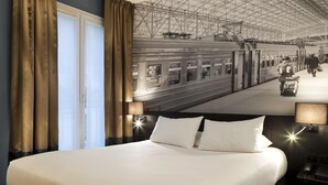 Habitación de hotel con una cama grande, una mesita de noche con lámpara y vistas a una estación de tren con un tren y gente.