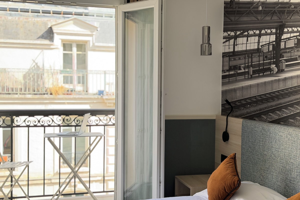A hotel room with a bed, a small table, a chair, and a balcony with a view of a building.