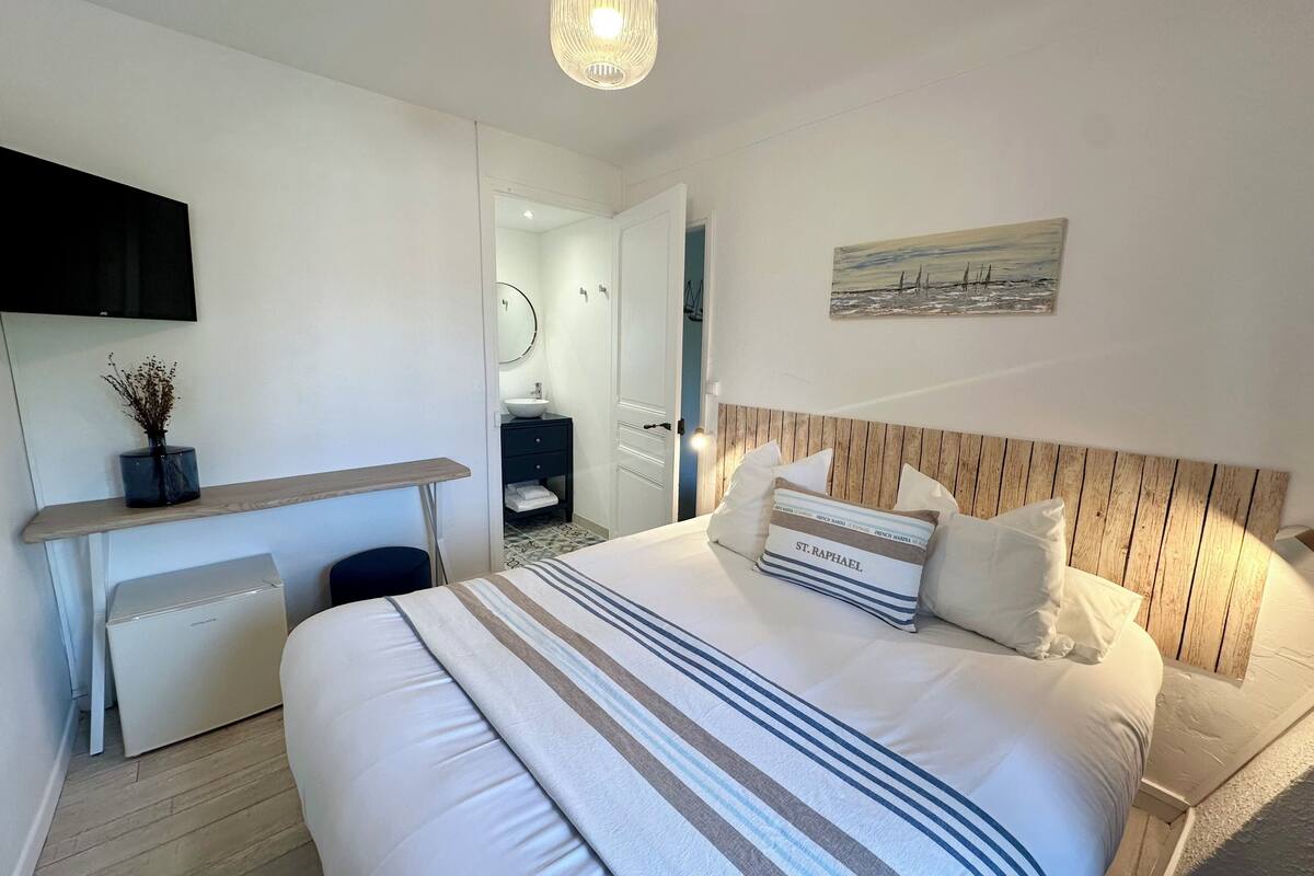 A hotel room with a bed, bedside table, a small desk, a mirror, and a painting on the wall.