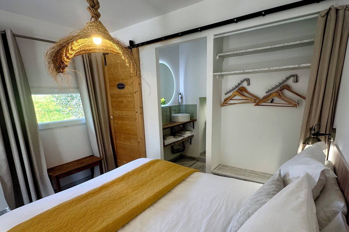 A hotel room with a bed, a hanging light fixture, a bathroom with a mirror, and a coat rack.