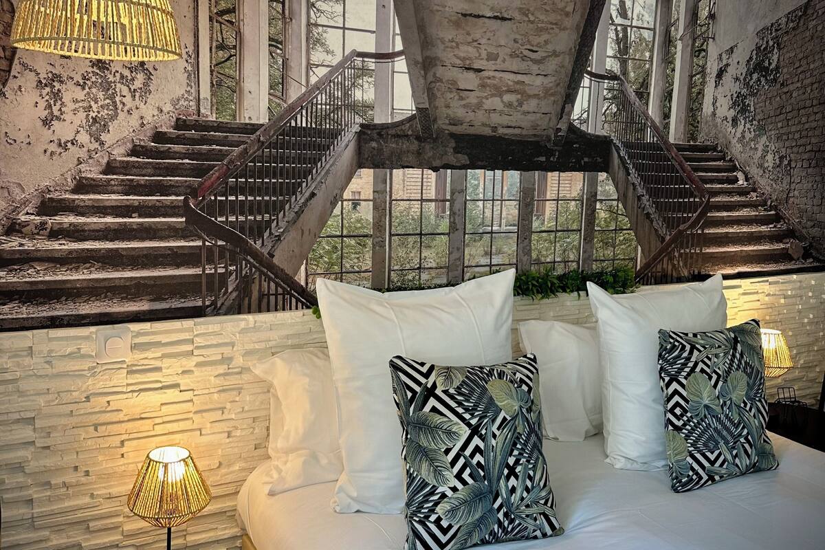 A bedroom with a bed, pillows, a nightstand, a lamp, and a staircase leading to an outdoor area.