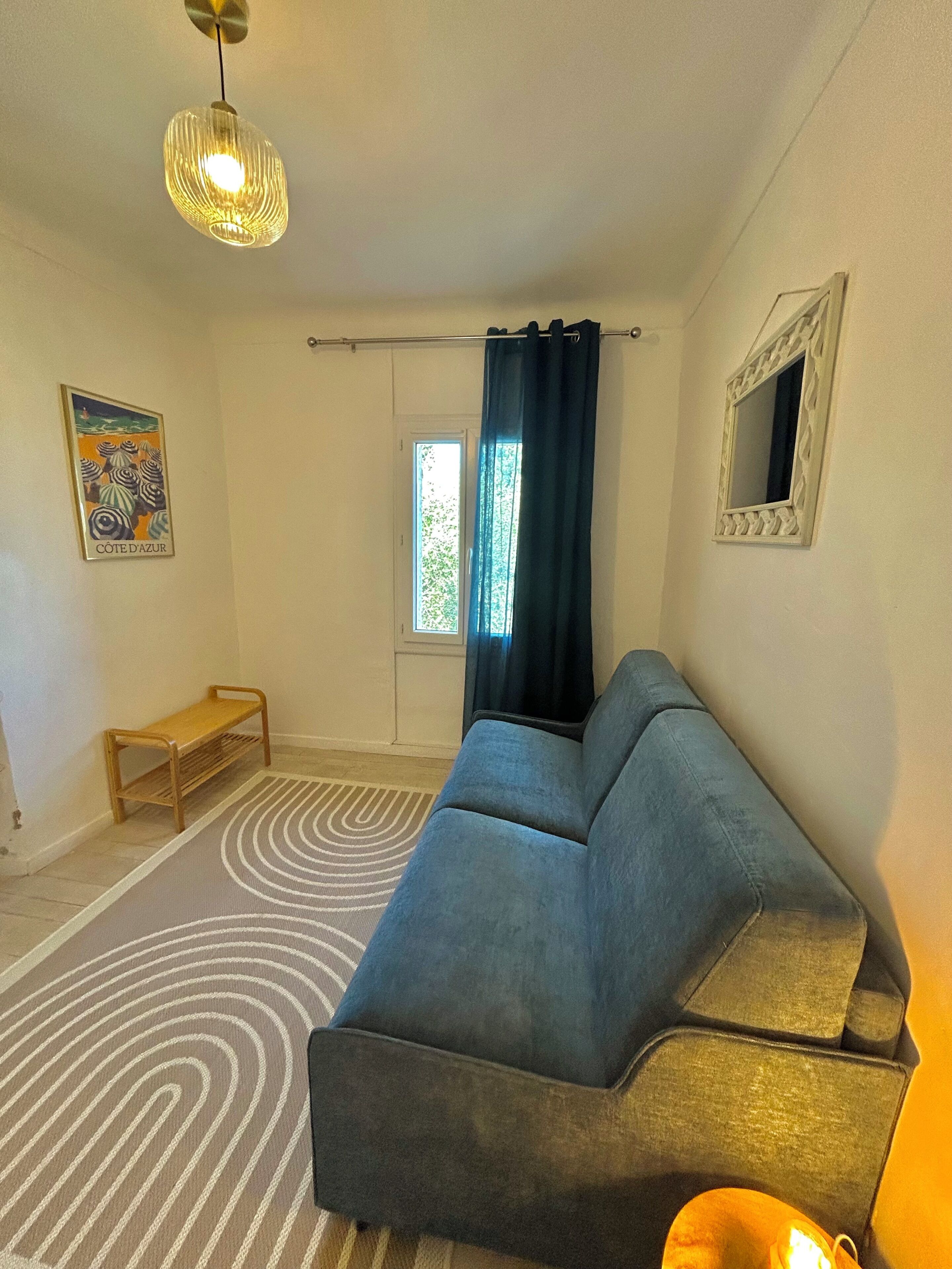 Una sala de estar con sofá, lámpara colgante, banqueta, alfombra estampada, ventana con cortinas e imagen en el marco en la pared.