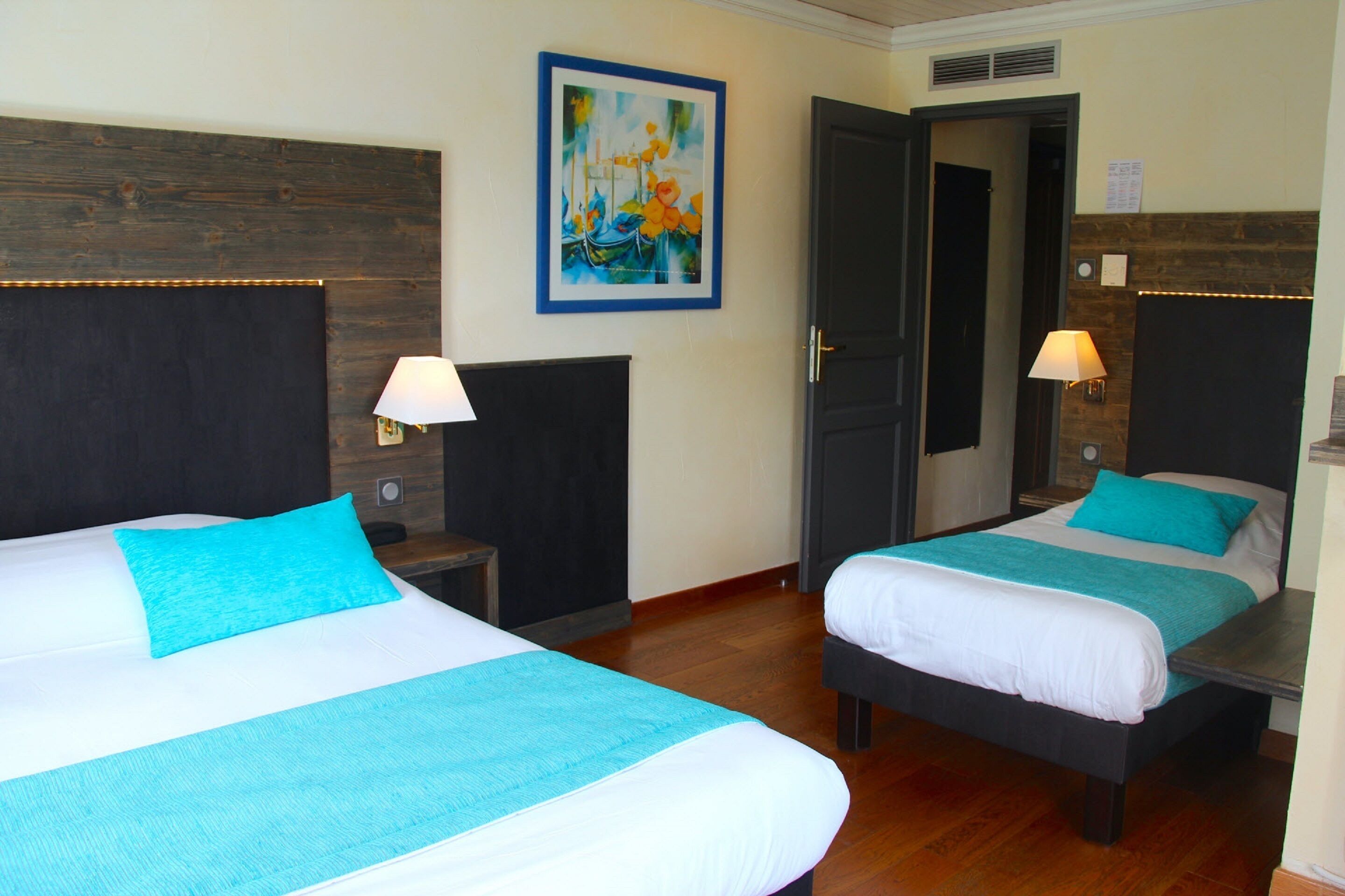 Dos camas individuales con ropa de cama azul y blanca a juego, un cabecero de madera, un cuadro en la pared y una puerta que da a otra habitación.