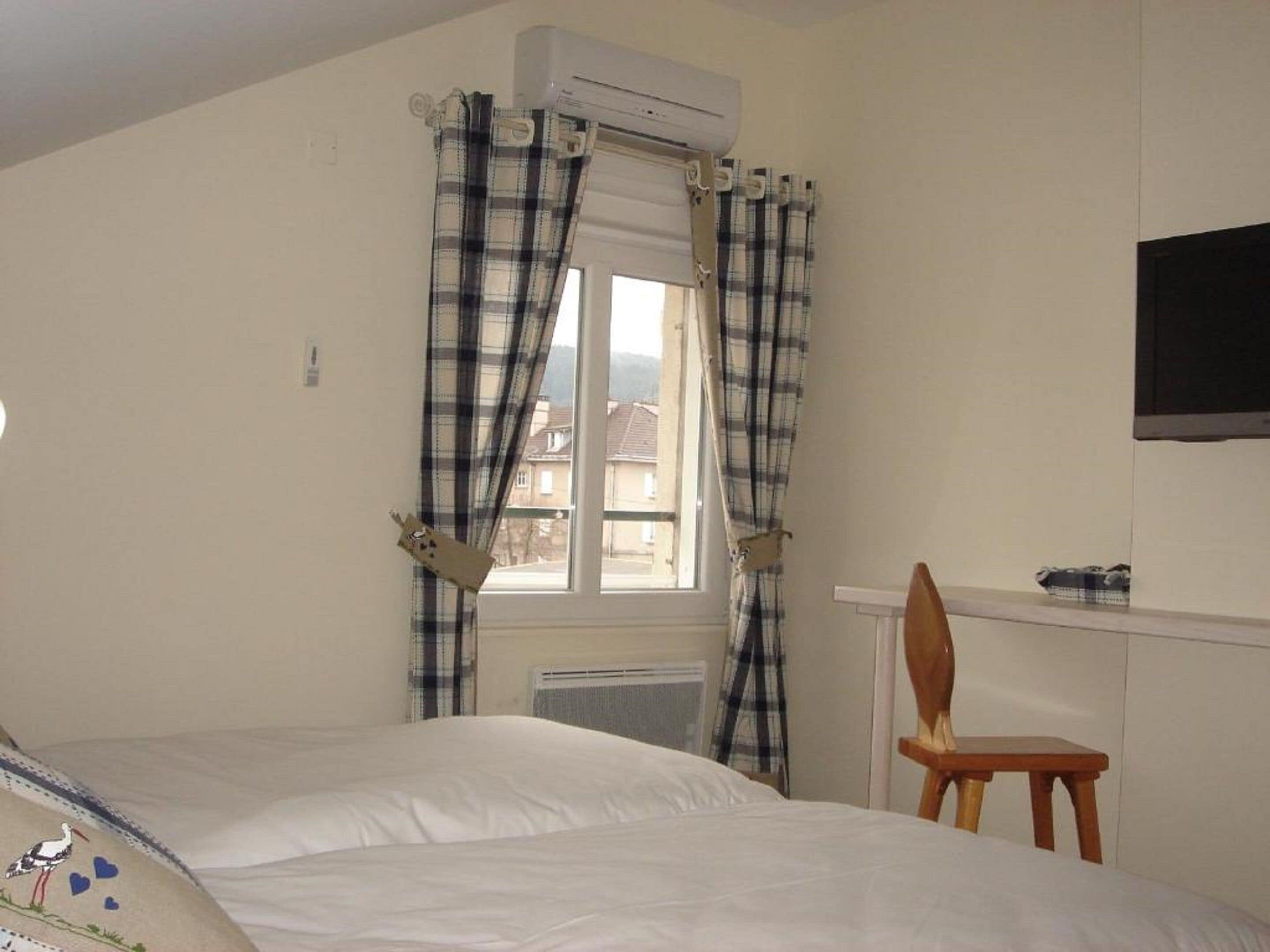 A bedroom with a bed, a chair, a window with curtains, an air conditioning unit, and a wall-mounted television.