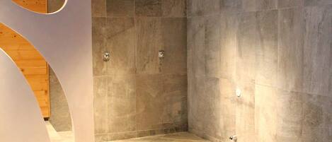 A modern shower with a stone floor and walls, a wooden panel on the left, and a recessed ceiling light.