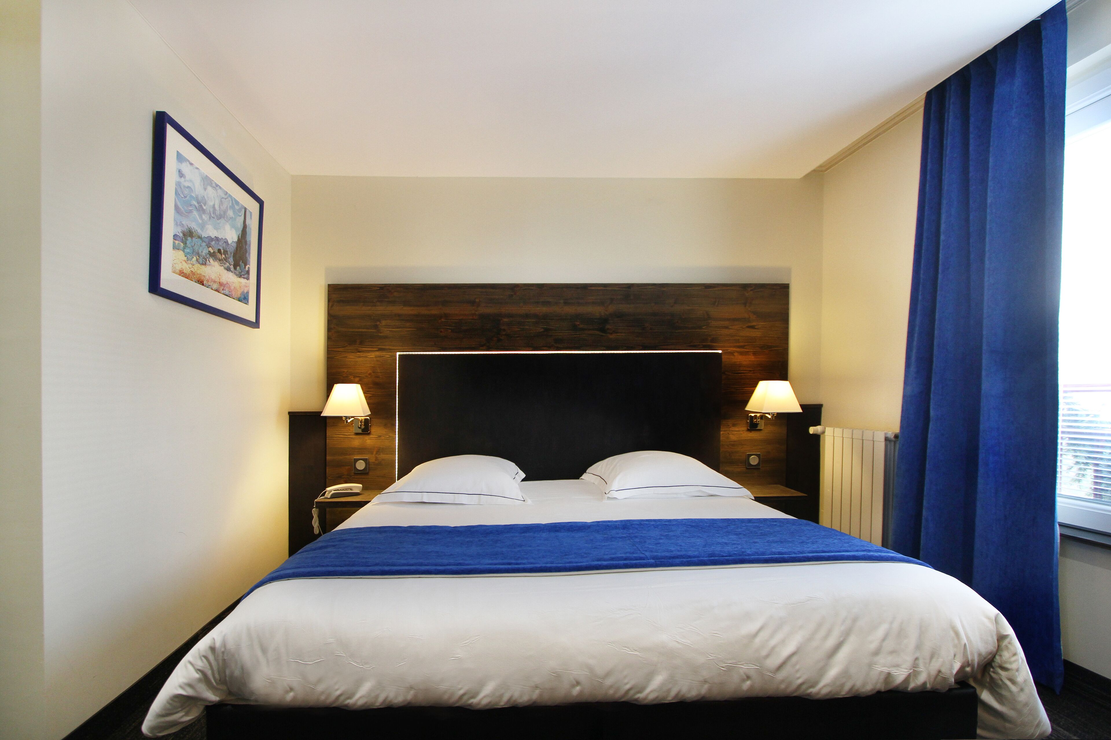 Hotelkamer met een groot bed, twee nachtkastjes met lampen, een houten hoofdbord, een ingelijst schilderij aan de muur en een raam met blauwe gordijnen.