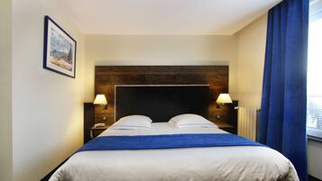 Hotelkamer met een groot bed, twee nachtkastjes met lampen, een houten hoofdbord, een ingelijst schilderij aan de muur en een raam met blauwe gordijnen.