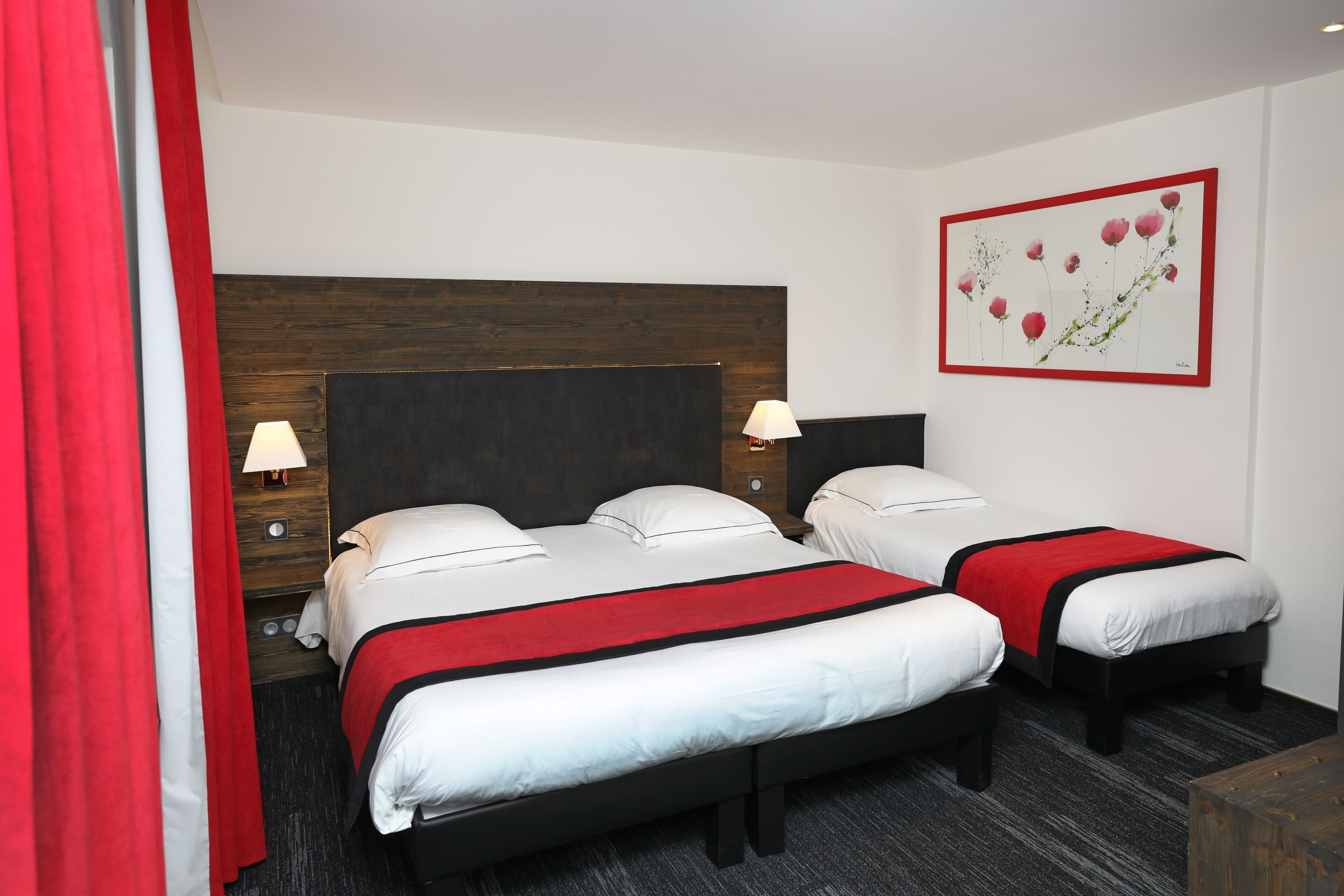 A hotel room with two beds, a wooden headboard, a red and white bedspread, a wall-mounted lamp, and a framed floral artwork.