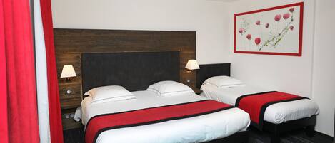 A hotel room with two beds, a wooden headboard, a red and white bedspread, a wall-mounted lamp, and a framed floral artwork.