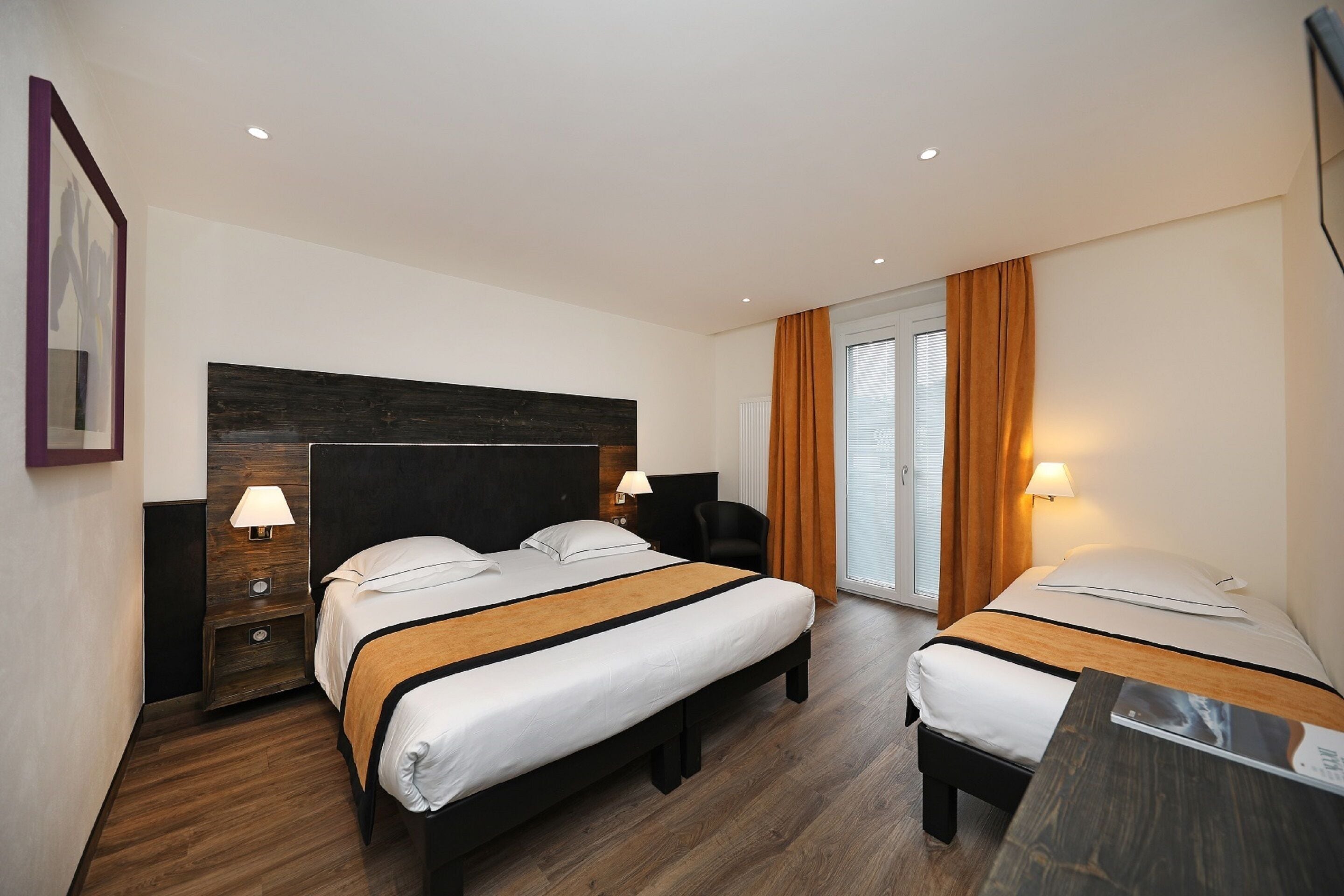 A hotel room with two beds, wooden flooring, a large dark headboard, and a sliding glass door with curtains.