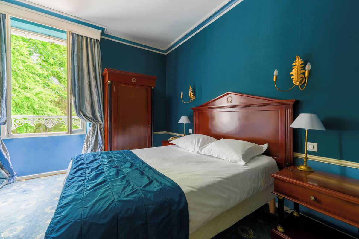 Een slaapkamer met een bed, nachtkastjes, een dressoir, een raam met gordijnen en uitzicht op groen.